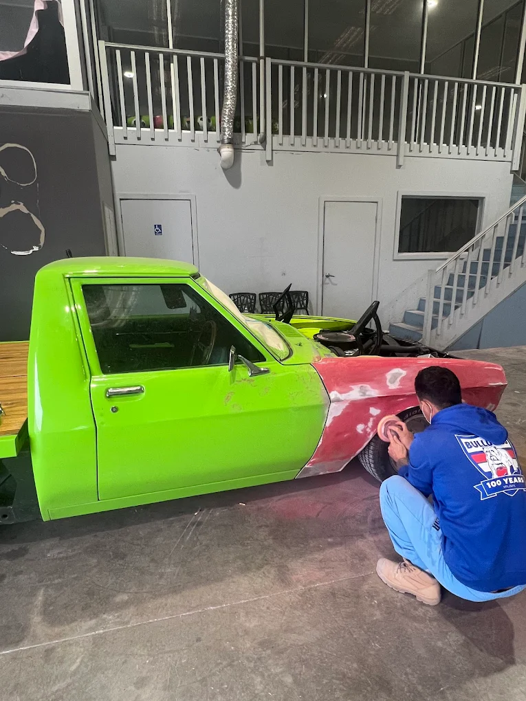 Technician hand-sanding front fender of classic car mid-restoration — red primer visible